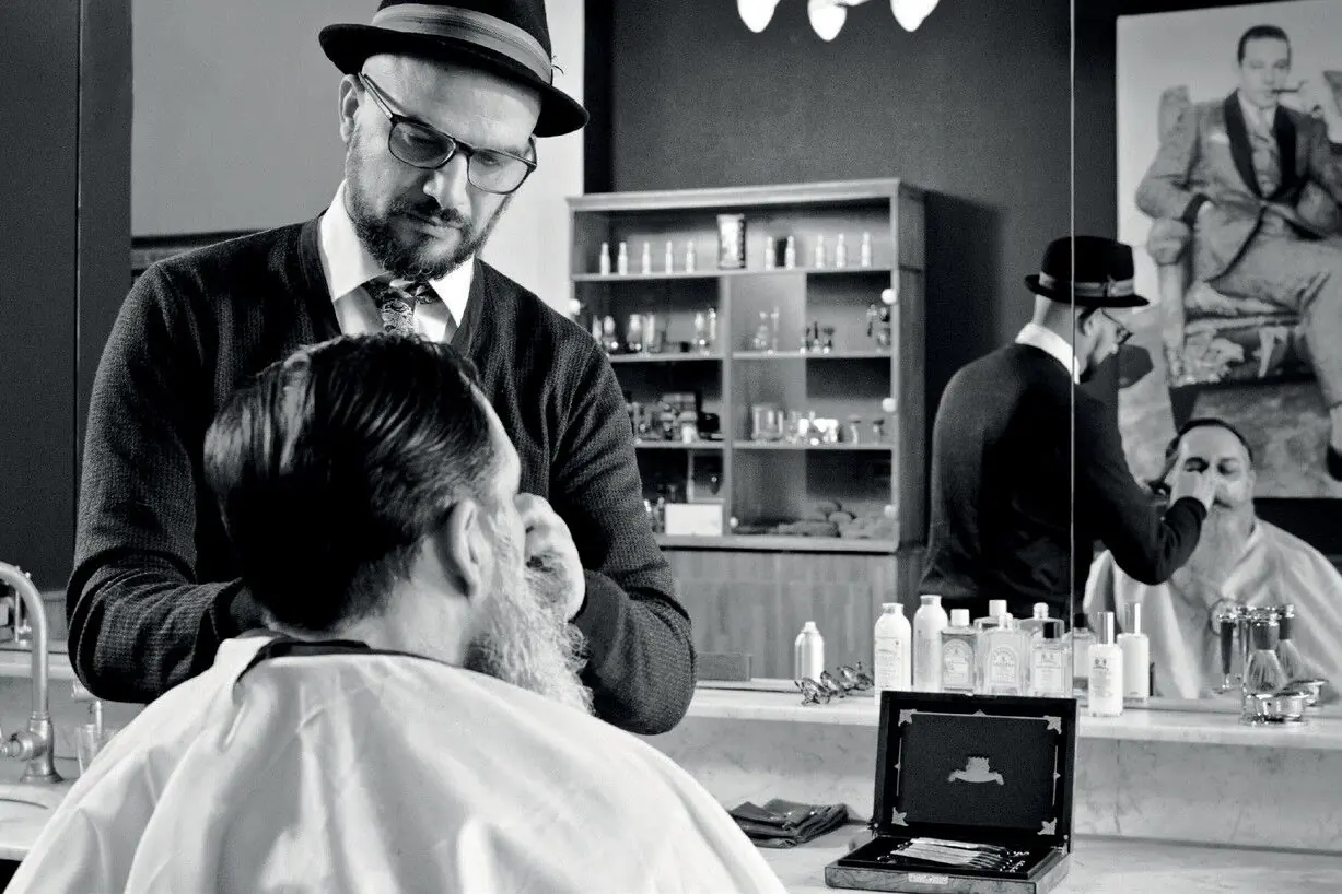Eddine Belaid arbeitet mit Hut und Brille am Bart eines Kunden im Barbershop, schwarzweiss