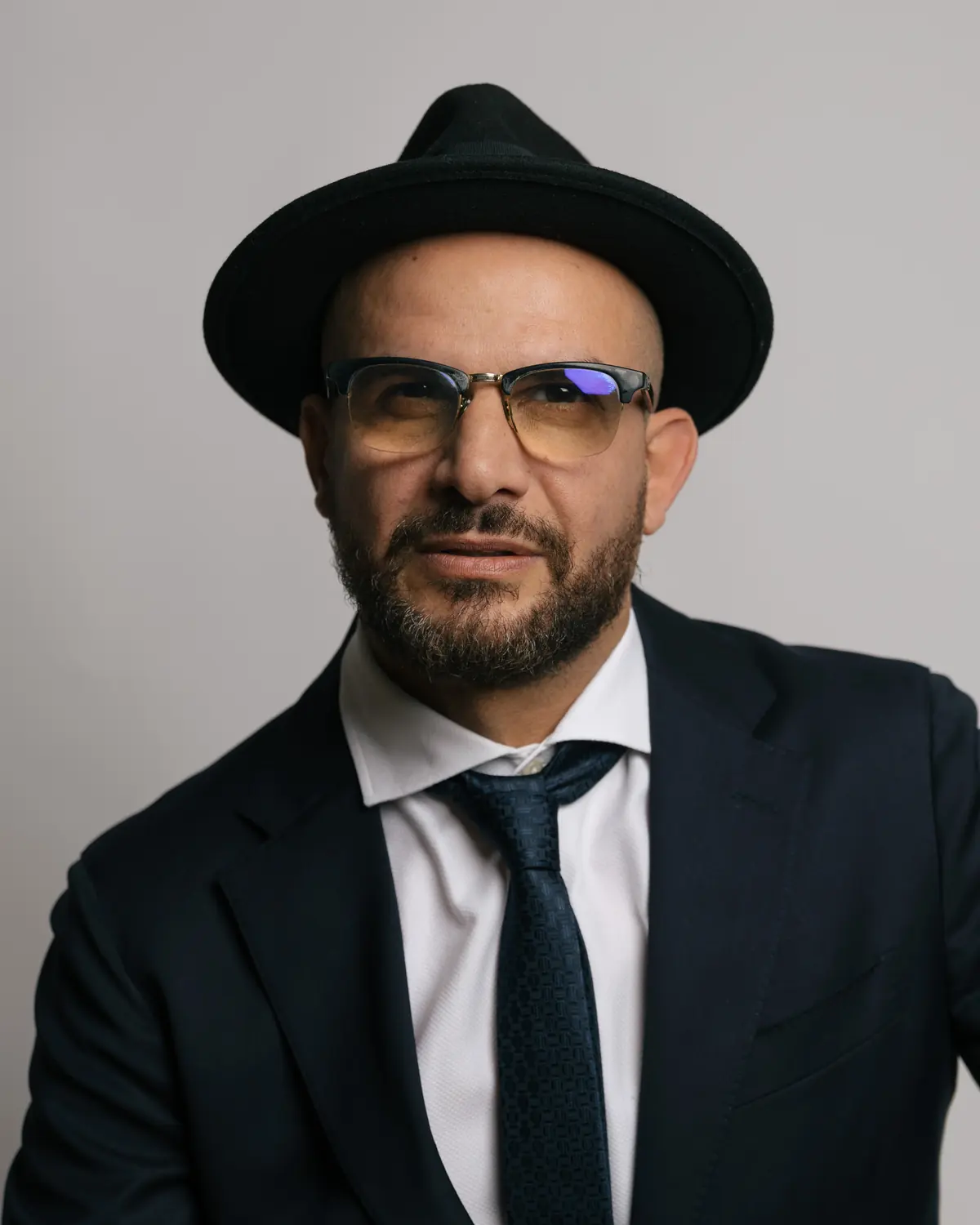 Eddine Belaid, Coiffeur und Barbier aus Zürich, mit Hut und Brille im dunklen Anzug vor hellgrauem Hintergrund